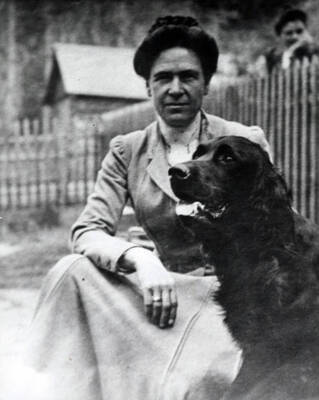 Charlotte Bovill posed seated with a dog beside a fence. In the background, a person is looking over the fence toward Mrs. Bovill.