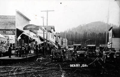 Book caption: On the left, in order, are the pool hall, post office; Peterson building (Albert Pierce store), and a building (previously the Olson store) which at this date house the barber shop and Dr. Faust's office. The other large bldgs. have already been identified. The large building above the bank was, at this time, occupied either by Olson or Sam Anderson. Of interest in this picture are the cars. The forward three of the line-up are a Reo owned by Jorgeson, a Winton owned by Drury, and a Studebaker. The REO is a 1911 REO 30 Touring car, capacity of 5 passengers. The Winton is probably a 1910 Winton Six (by the number of passengers that appear, the removal of the roof makes the identification difficult). The Studebaker is believed to be a Garford, but may be unidentifiable based on this photograph.
