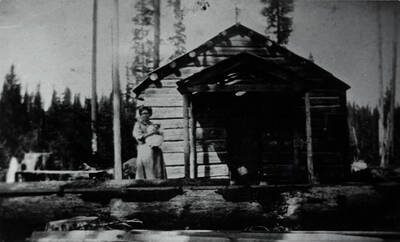 A one-room log school house at Collins, Idaho. Mrs. Crawford, the teacher, stands next to the school.