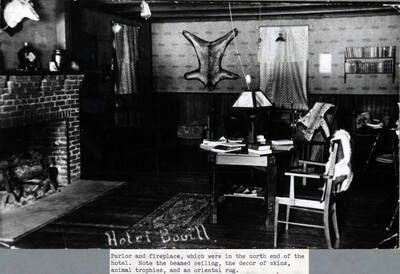 A photograph of the interior of the Bovill Hotel parlor and fireplace, located in the north end of the building. Note the beamed ceiling and the décor of skins, animal trophies, and an oriental rug.