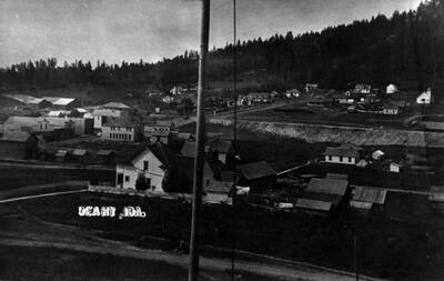 Buildings are identified in book caption: 'Looking northwest from a point not far from the school. The McQueary Hotel hides part of the west side of Main Street. Above it are the hardware and drug store, both characterized by false fronts with peaks. The flat roof between is part of the drugstore. Next is the box-like shape of the bank. Up the street, partly hidden, is a larger building that at this time or later housed the Olson store, and over is is seen the roof of the depot. The largest building on the east side of the street - the one with many windows - is the Carlson Hotel. Above it, just below the railroad crossing, is the newspaper office.'