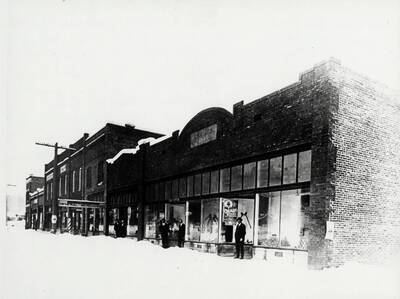 The new buildings of Main Street in January 1915. Just after reconstruction of the town. (The plan here conforms to what is shown on Map 5 in the book 'The Trees Grew Tall,' by John B. Miller). Seen in the first doorway is E.K. Parker. In the 2nd, with another man a little to one side, is Dan Featherstone. A showbill: 'Black Crow' minstrel type - The Oldest and Best Theatre - Tuesday, January 12.' Another bill on the wall of the store is readable in except for three words here reproduced as blanks: The Road to Ruin - an uplift drama with a cast of Broadway stars. The _____ _____ _____ people - 250 scenes.'