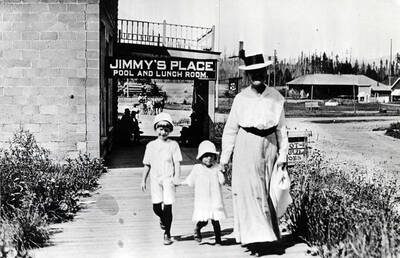 A woman and two children walk down the sidewalk in Bovill. From the caption in Trees Grew Tall: 'The view past the Verdon premises toward the depot. At this stage, the concrete building was occupied by Jimmy Gilroy.'