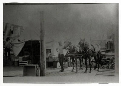 John Groh (standing by horses) operated his first grocery store out of a tent. The man at left is not identified. Beside him are three large squash; elsewhere are crates of vegetables and melons. In the background at left is the frame of a new building, probably the Gropp store. Dimly seen above the horse's rump is a two-story building once the J.A. Anderson store, which stood at Pine and 1st.