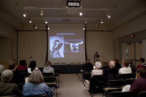 Photograph  6 of Robert Caisley, Marco Deyasi, and Lauren 'Lojo' Simon's Colloquium Panel Discussion 'Art about Art: Collaboration and Expression of Self and Other in Creating New Work for Theatre (on the play "Adoration of Dora").' Robert Caisley is Associate Professor of Theatre Arts and Head of Dramatic Writing. Marco Deyasi is Assistant Professor of Art and Design. Lauren 'Lojo' Simon is a graduate student. Pictured at podium: Marco Deyasi.