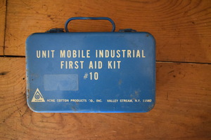 A blue metal box. "Unit Mobile Industrial First Aid Kit #10, Acme Cotton Products Co., Inc. Valley Stream, N.Y. 11582" is printed on the box.