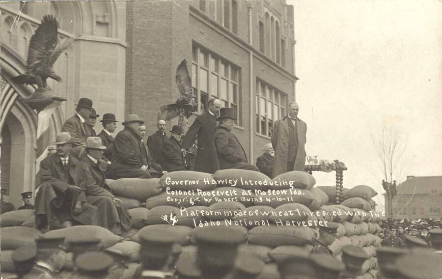 Former president, Colonel Theodore Roosevelt, sits on a platform in front of the University of Idaho Administration Building during his visit in 1911. Writing on image: 'Governor Hawley introducing Colonel Roosevelt at Moscow Ida. Copyright by Robert Burns 4-10-11. Platform made of wheat threshed with the Idaho National Harvester #4'.