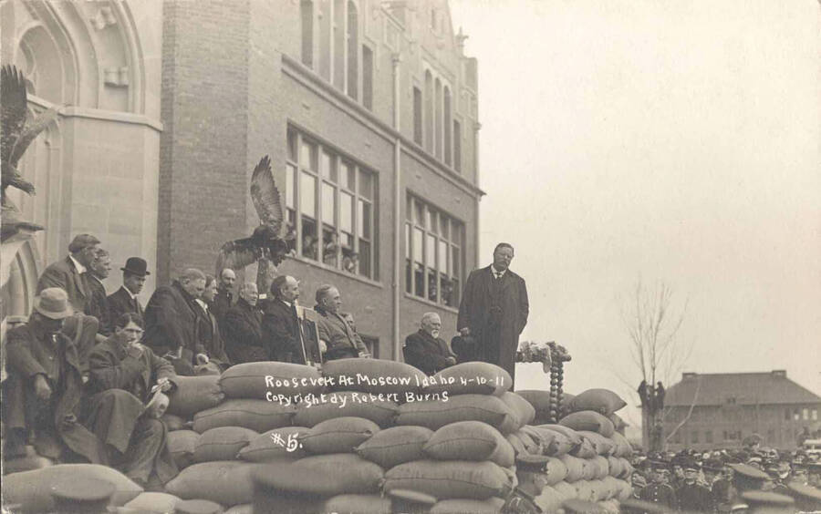 Theodore Roosevelt speaking on platform in front of University of Idaho Administration Building. Writing on image reads: 'Roosevelt at Moscow Idaho 4-10-11 Copyright by Robert Burns #5.'