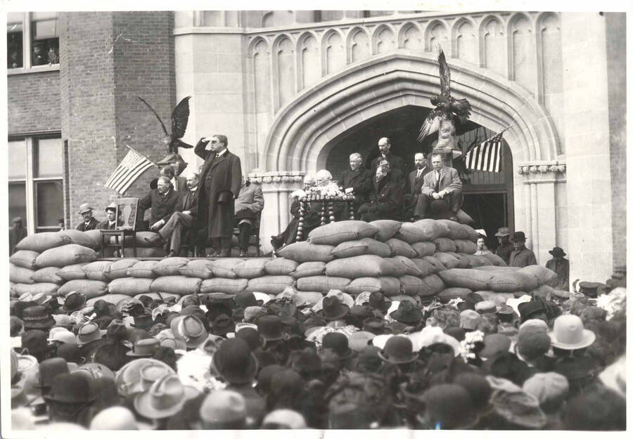Theodore Roosevelt speaking on platform in front of University of Idaho Administration Building. He is shielding his eyes.