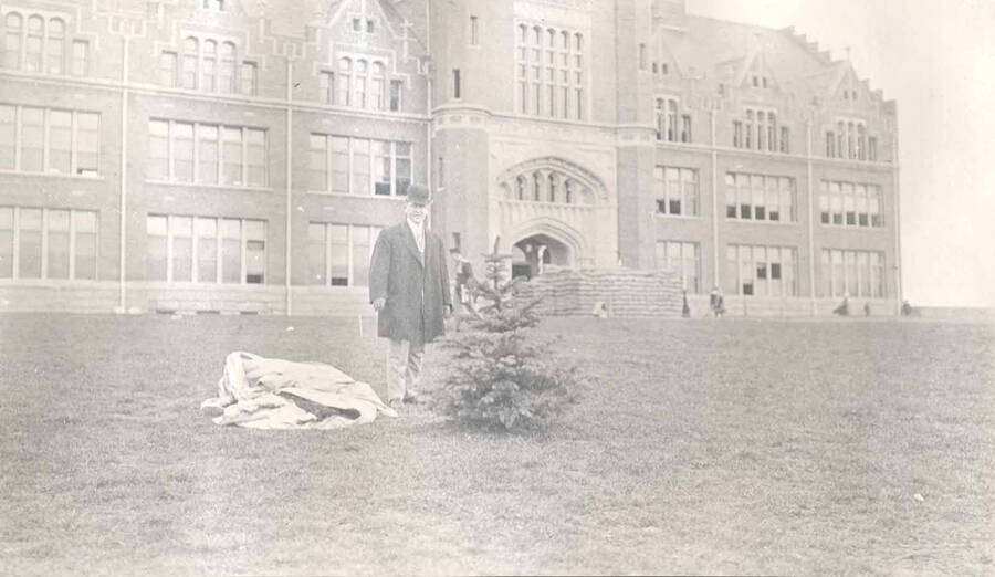 An unidentified man stands next to a small tree planted by Theodore Roosevelt in front of the University of Idaho Administration Building.