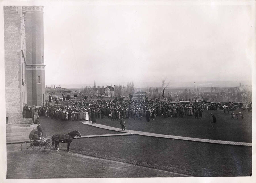 A crowd assembles in front of the University of Idaho Administration Building to hear Roosevelt speak. Boardwalk leading to Administration Building is visible in foreground, as is a horse and buggy on the left. 