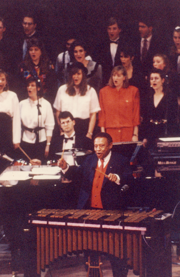 A person holding mallets in front of a vibraphone with musicians behind him and rows of singers in the background.