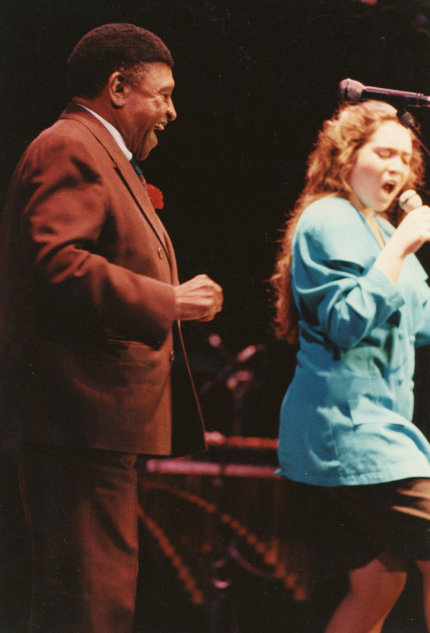 A person watching another sing into a microphone with the blurred outline of keyboards in the background.