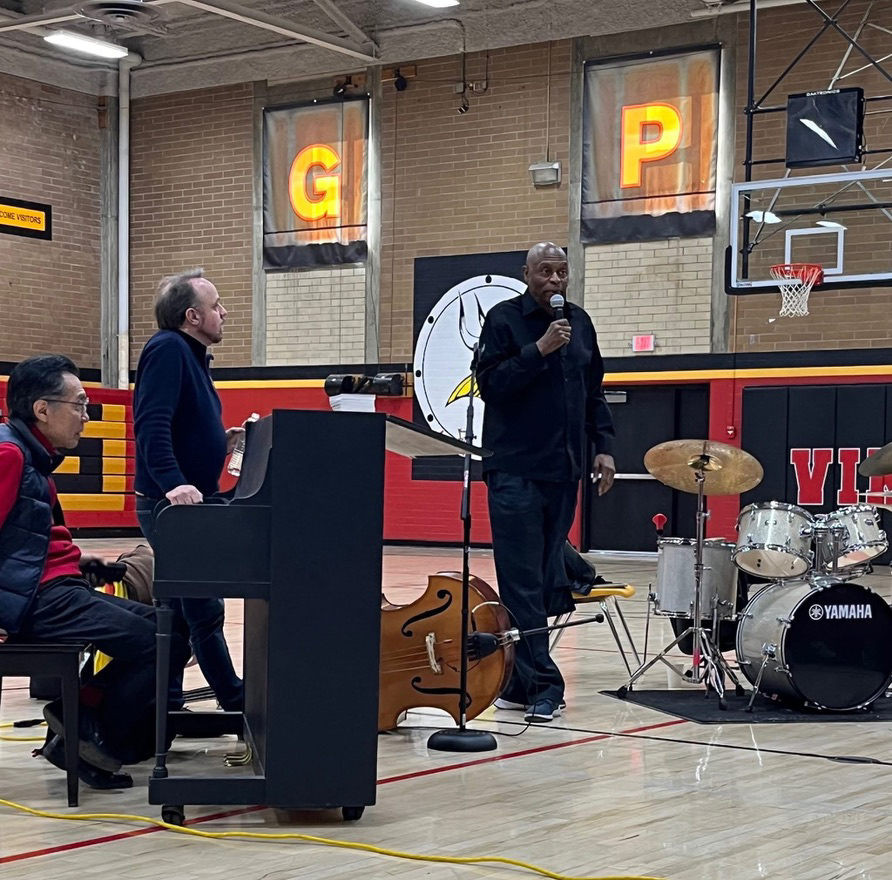 A person playing piano and another standing by it watch a person in front of them speak into a microphone, with a double bass and drum set beside him. They are in a gymnasium with the letters G and P hanging from the wall.