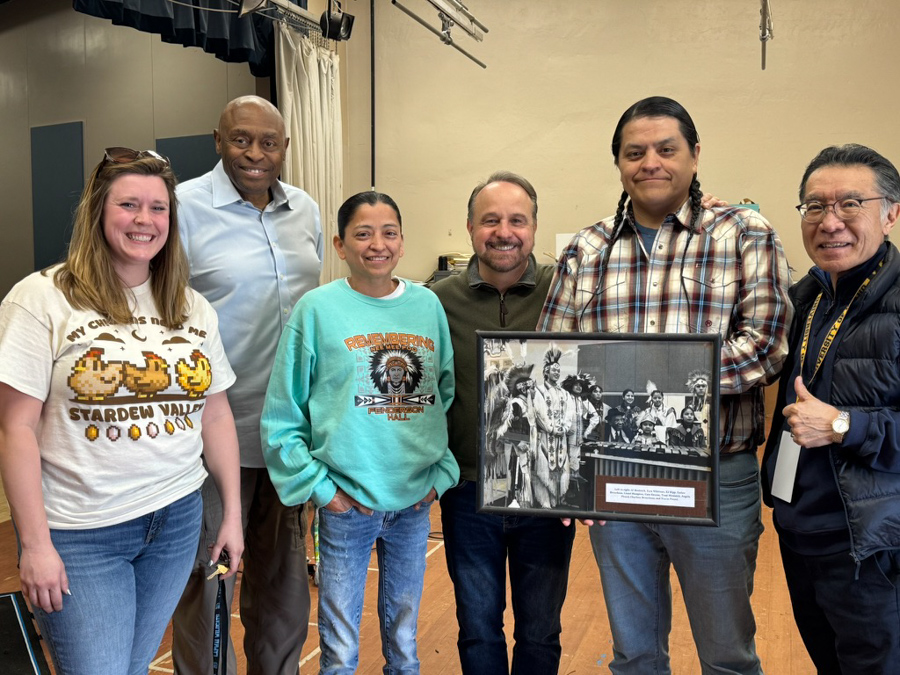 A group of six people pose with a black and white historic photo featuring another group in traditional Indigenous dress.