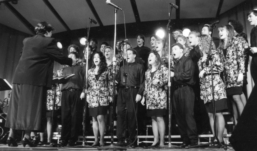 A conductor leads three rows of singers with four stands of microphones in the foreground and stagge lights visible in the background.