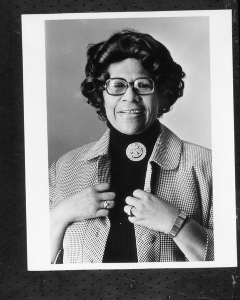 A portrait of a person smiling, wearing a checkered jacket, a black turtleneck sweater and a brooch with the design of a smiling face.