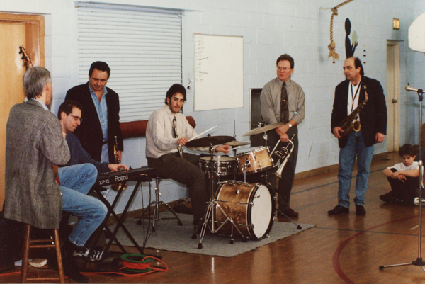 A group of six people sit and stand as they either play or hold jazz oriented instruments in a practice space with cement walls. A child sits on the right side of the frame.