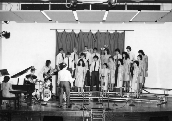 A pianist, drummer and bass player on the left side of the image support three rows of singers performing in front of a banner, being led by a conductor.