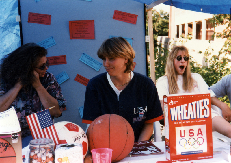 Lindy Wellyn and Andrea Lloyd at the Women's Center table at Palousafest. There is an American flag, soccer ball, basketball, and box of Wheaties on the table in front of them.
