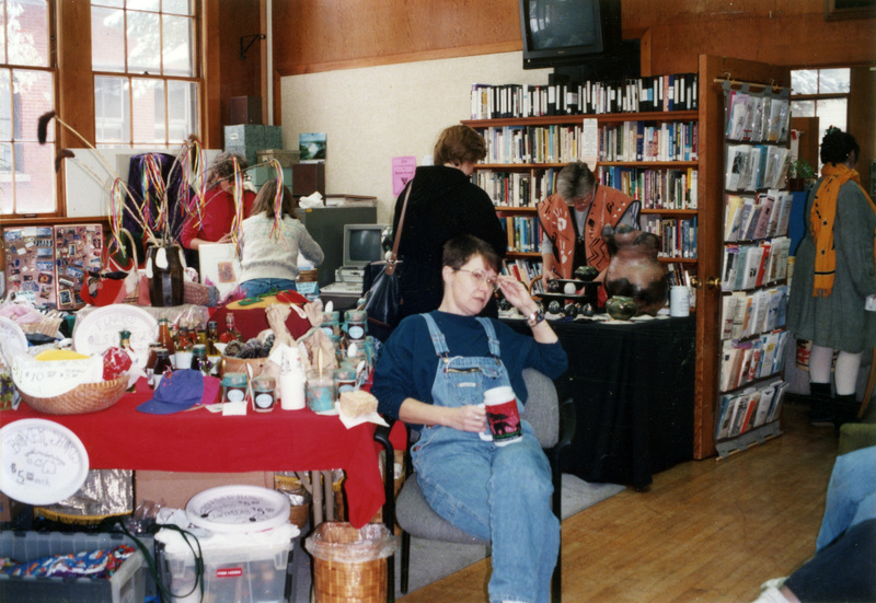 Gwen Snow, Rebecca Rod, and Jenny Rod in the Women's Center. One woman sits next to a table covered in items while another stands in front of a full bookshelf. Several other women are nearby.