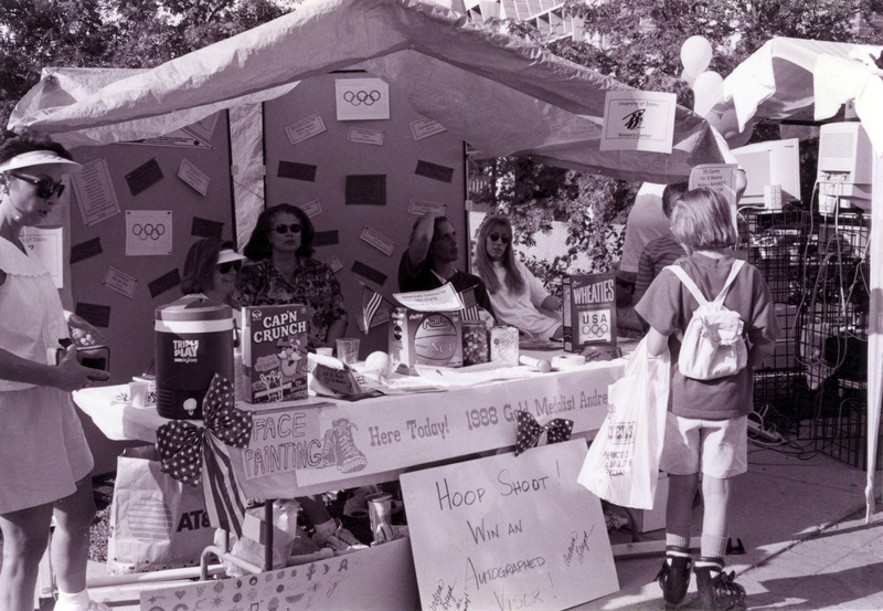 Val Russo (left), Jill Anderson, Lindy Wellyn, and Andrea Lloyd (far right) sit at a booth advertising the presence of Andrea Lloyd, a 1988 Gold Medalist, at the Women's Center Palousafest Booth. The booth also advertises face painting and a hoop shoot.