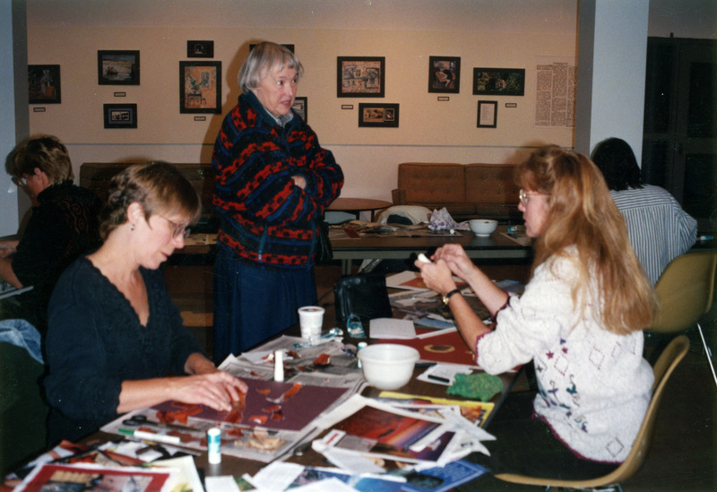 Pam Palmer, Janet Lecompte, and Mary Blythe sit at and stand near a table covered in crafting supplies. Two appear to be working on collages.