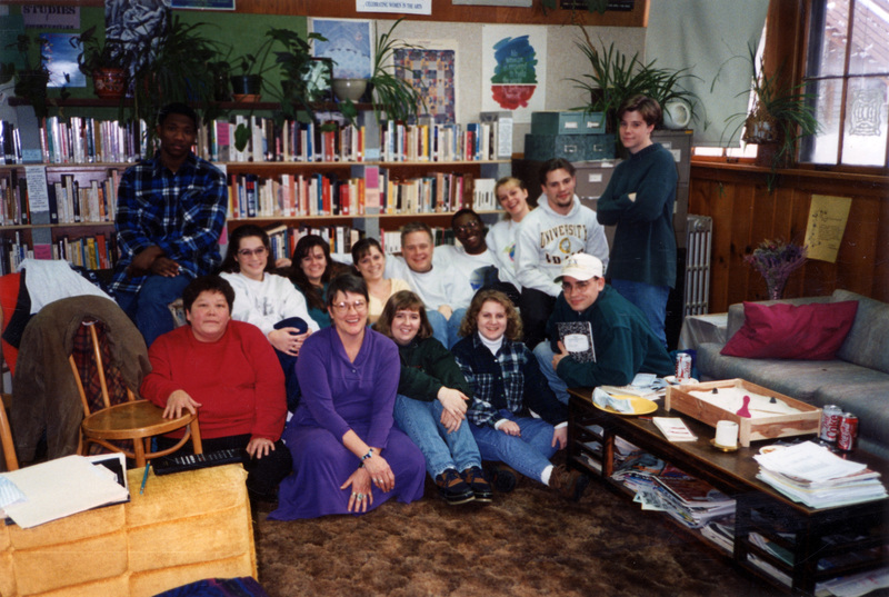 Betsy Thomas, the woman in purple, sits in a group portrait shot. The group is sitting on a brown carpet in front of a bookshelf.