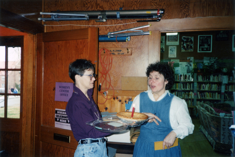 Gwen Snow (left) and an unidentified woman talking in the Women's Center Office. Gwen Snow is holding a pie with a lit candle in it.