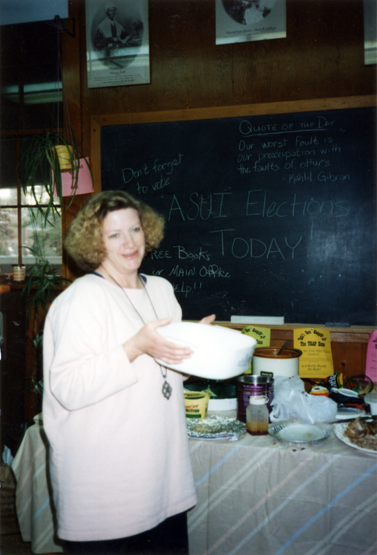 Jill Anderson standing in front of a blackboard in the Women's Center. The woman is holding a casserole dish. The blackboard reads "ASUI Elections Today! Don't forget to vote. Free books in Main Office [blocked]elp!! Quote of the Day: "Our worst fault is our preoccupation with the faults of others." - Kahlil Gibson."