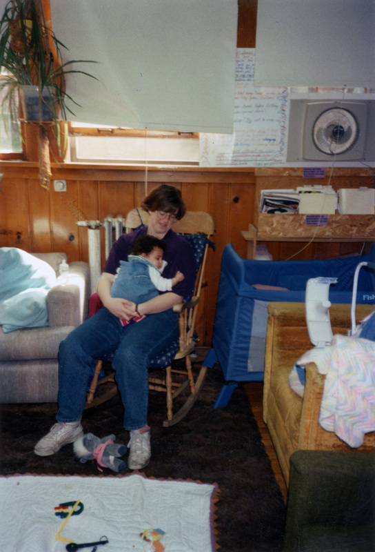 Christine and Jordan sitting in a rocking chair in the Women's Center in spring 1994. Baby toys are spread out on a blanket on the floor.