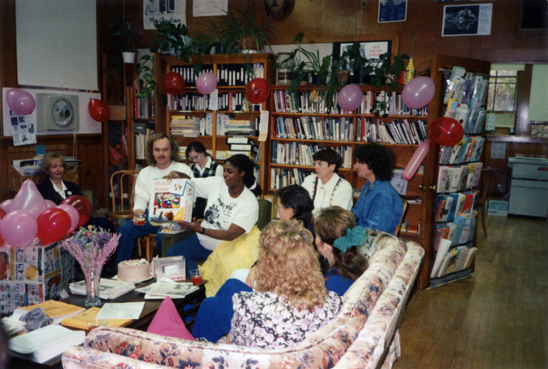 A baby shower for Women's Center secretary Leslie Gregory in fall 1993. She's holding a Musical Mobile and smiling. The room is covered in pink and red balloons.