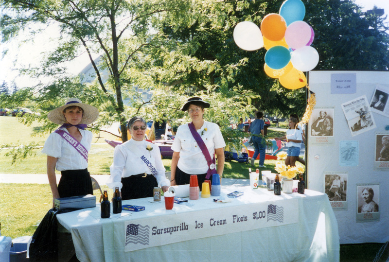 Three women standing at an outdoor table advertising Sarsaparilla Ice Cream Floats for $1. All three are wearing purple and white sashes. One reads"Women's Equality," while another reads "March for [blocked]."