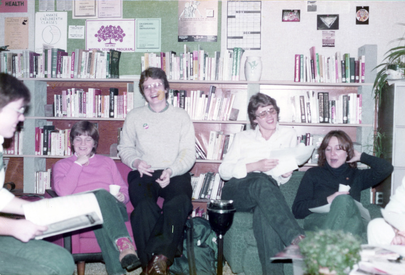 A group of women laughing in the Women's Center. Carolyn Caster sits in the middle of the five, third from the left. Two appear to be looking through pamphlets.