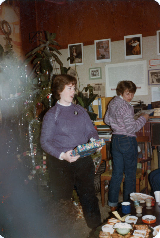 A person holds a present during a Christmas celebration at the Women's Center. There's a tree covered in tinsel and paper chains in the background. The table in the foreground is laden with snacks.