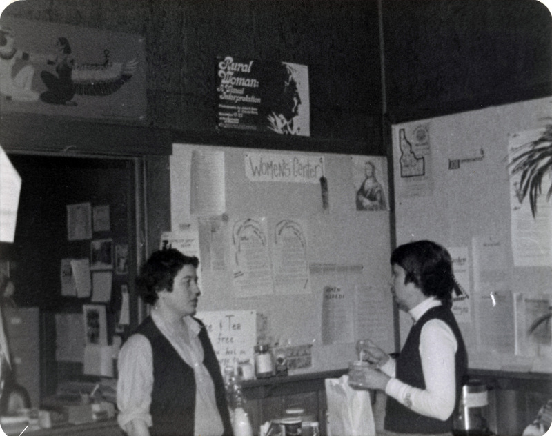 Left to right: Unidentified woman and Alayne standing in the Women's Center. They appear to be talking.