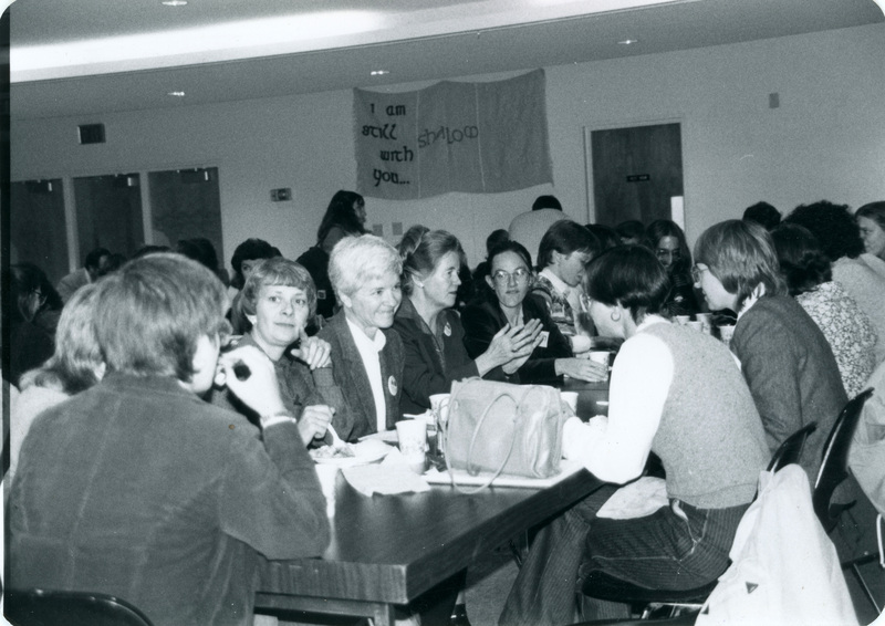 A large group of people meeting together at the potluck. Alayne Pettyjohn looks toward the camera, while Kay Keskinen sits across the table, second from the left. Peggy McIntosh is in the center of the shot with her hands raised.