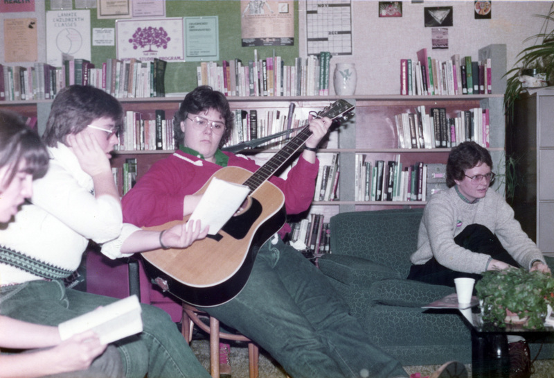 A person playing guitar at an event in the Women's Center. Two people are holding books.