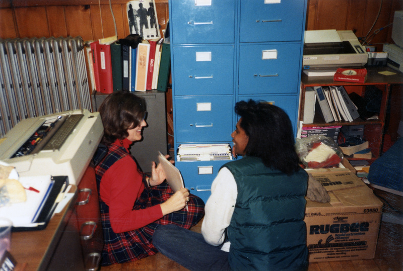 Betsy Thomas and Michelle Ward in the Women's Center office filing things into a filing cabinet. They're sitting on the floor and appear to be laughing about something.
