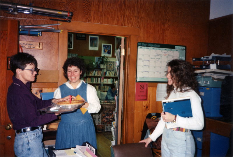 Three women standing in the Women's Center. From left to right: Gwen Snow, Kim Bouchard, and unidentified woman. Snow is holding a pie with a candle stuck in it.