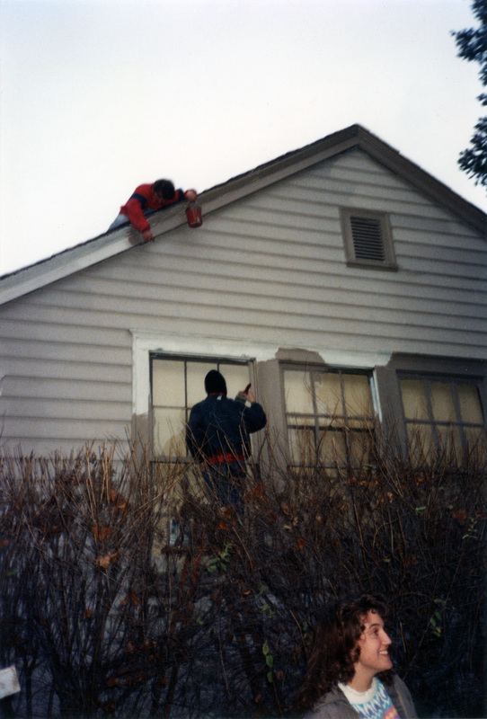 Two people painting a building. One is lying on the roof and pointing, while the other holds a paintbrush and stands near the windows.