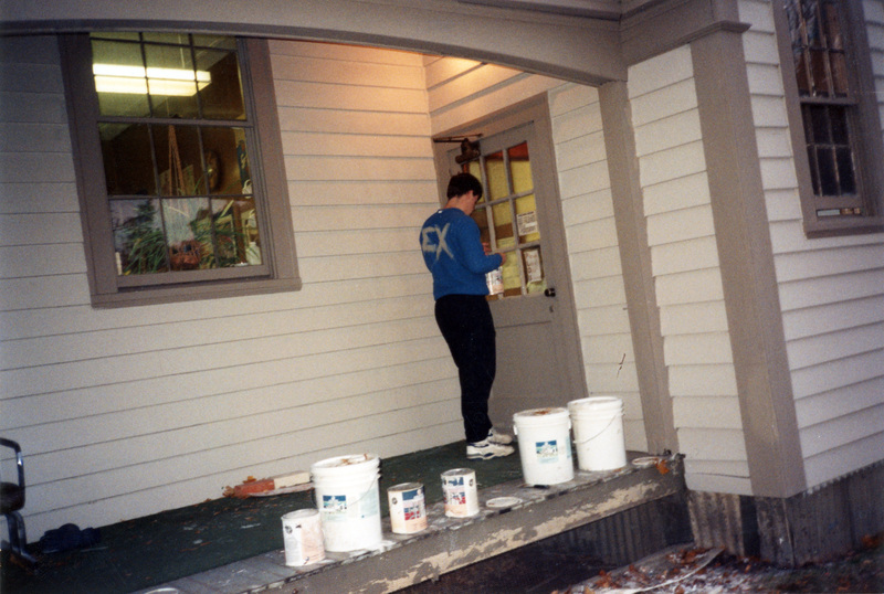 A person stands near a door and working on the Women's Center building. Several large buckets of paint are lined up along the edge of the porch.