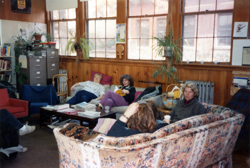 Several women, including Jenny Rod, sitting in the Women's Center during the Thanksgiving Potluck. Jenny Rod is the woman sitting on the couch underneath the window, wearing purple pants and looking at a book with a yellow jacket. 