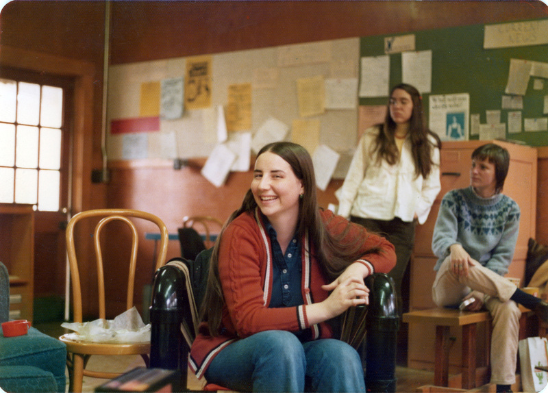 Hope Hadly smiles at the camera while two women stand and sit in the background inside the Women's Center. Hope Hadly is sitting on a custom chair made by Trynn Spiesman. The woman on the right in a grey sweater is sitting on a bench carved by Jeanne Wood with an image of Wonder Woman.