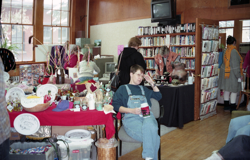 Gwen Snow, Rebecca Rod, and Jenny Rod in the Women's Center. One woman sits next to a table covered in items while another stands in front of a full bookshelf. Several other women are nearby.