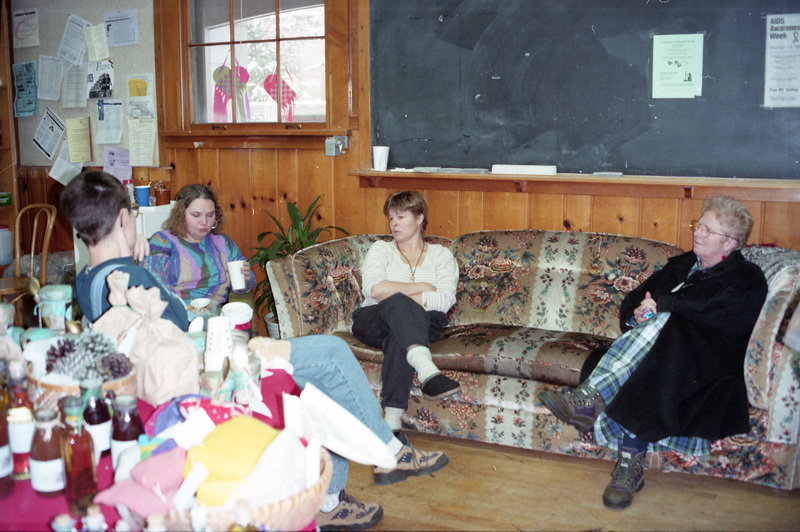 From left to right, Gwen Snow, Jeanne Wood, Gerri Sayler, and Ruby Valentine sitting in the Women's Center. Gerri Sayler and Ruby Valentine sit on a patterned couch underneath a blackboard. Gwen Snow and Jeanne Wood sit on separate chairs. A table filled with food and other objects is in the foreground on the left.