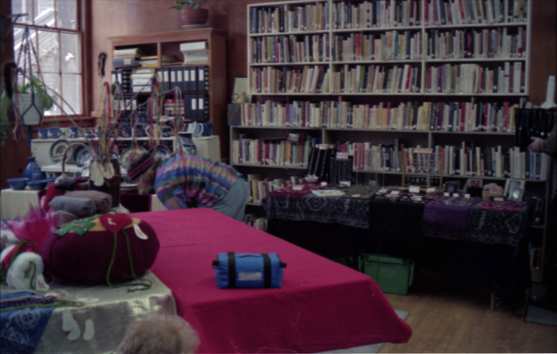 A shot of the inside of the Women's Center. A row of white bookshelves filled with books line the back wall. A table covered in bright magenta cloth is in the foreground. A person wearing bright striped clothing can be seen bending over behind the table.