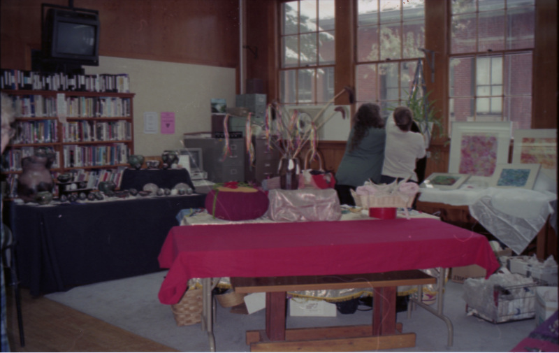 A shot of the inside of the Women's Center. A table covered in bright magenta is in the foreground. A television screen hangs off the wall on the left. Two women can be seen near large windows on the right.