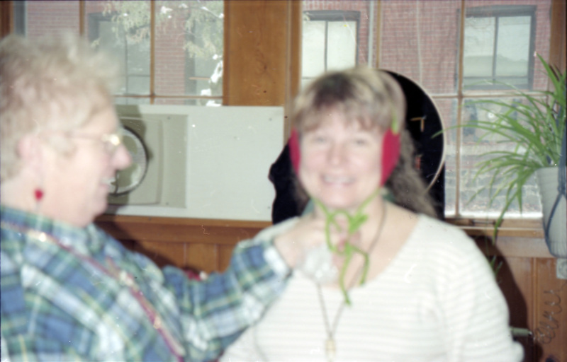 Ruby Valentine and Gerri Sayler sitting in the Women's Center. Gerri Sayler is wearing what looks like bright pink earmuffs. Ruby Valentine with her profile facing the camera and is holding onto green strings attached to Gerri Sayler's ear piece. The image is focused on the windows in the background.