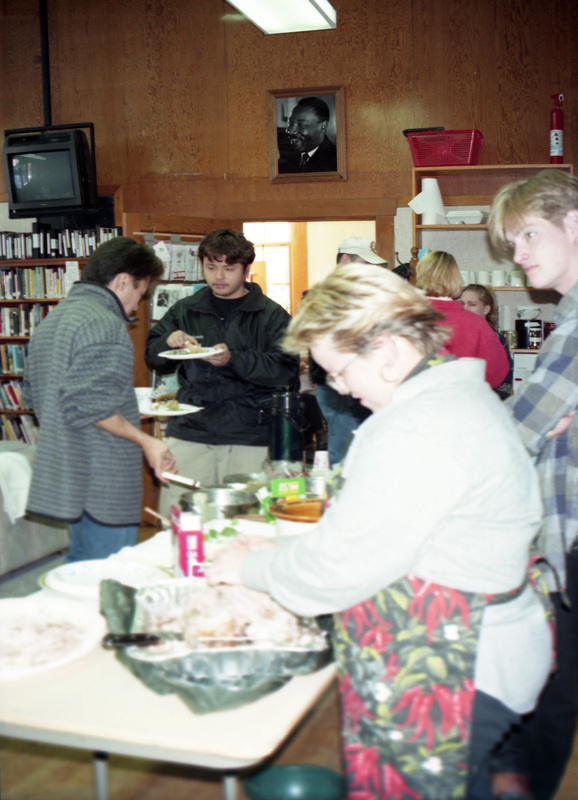 People serving and eating food in the Women's Center. A woman wearing a patterned apron stands over a foil tray in the foreground. A portrait of Martin Luther King Jr. hangs over the doorway in the background.